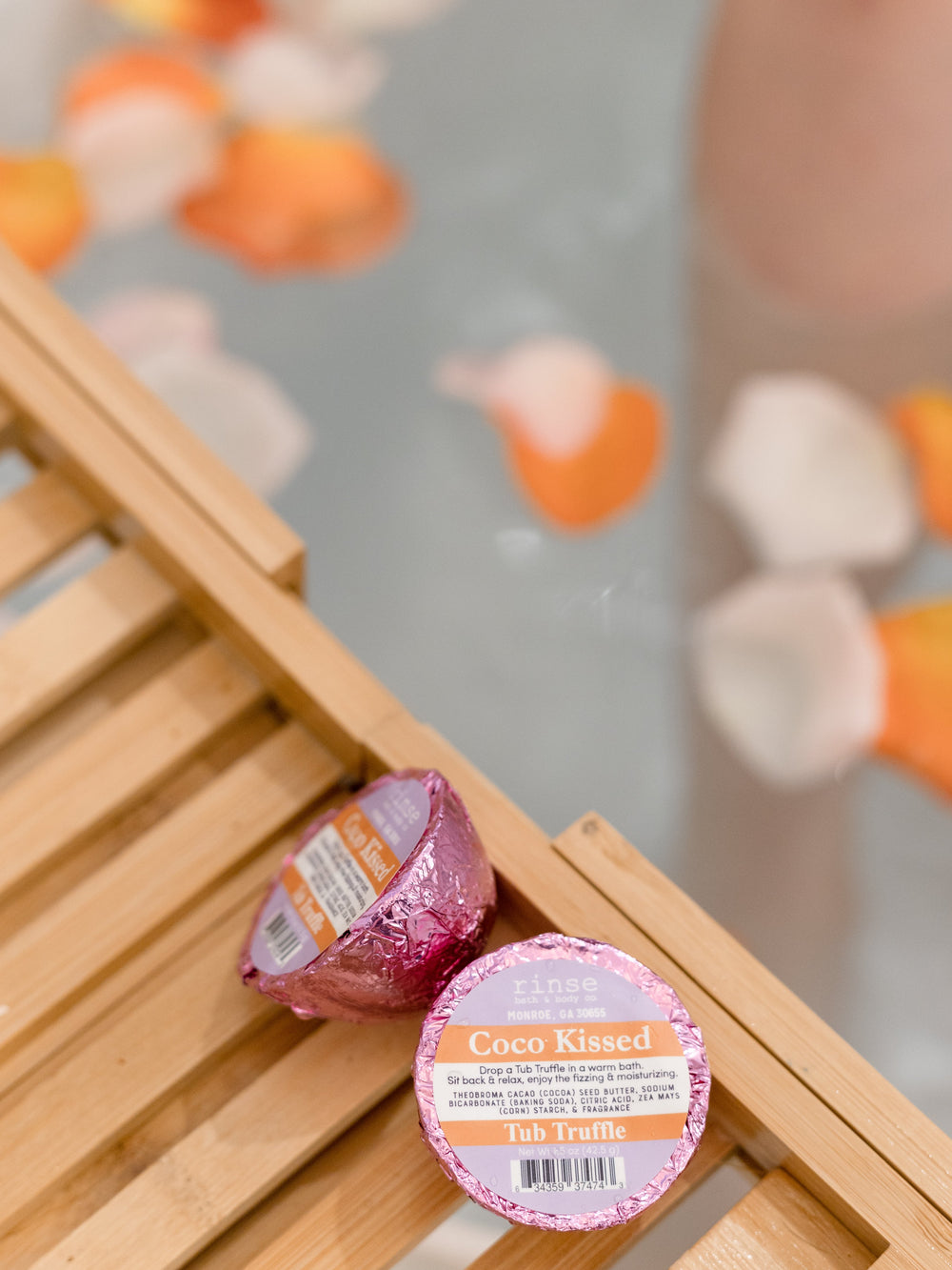 Two Coco Kissed Tub Truffles resting on a wood bath caddy — rose petals and submerged leg soft behind