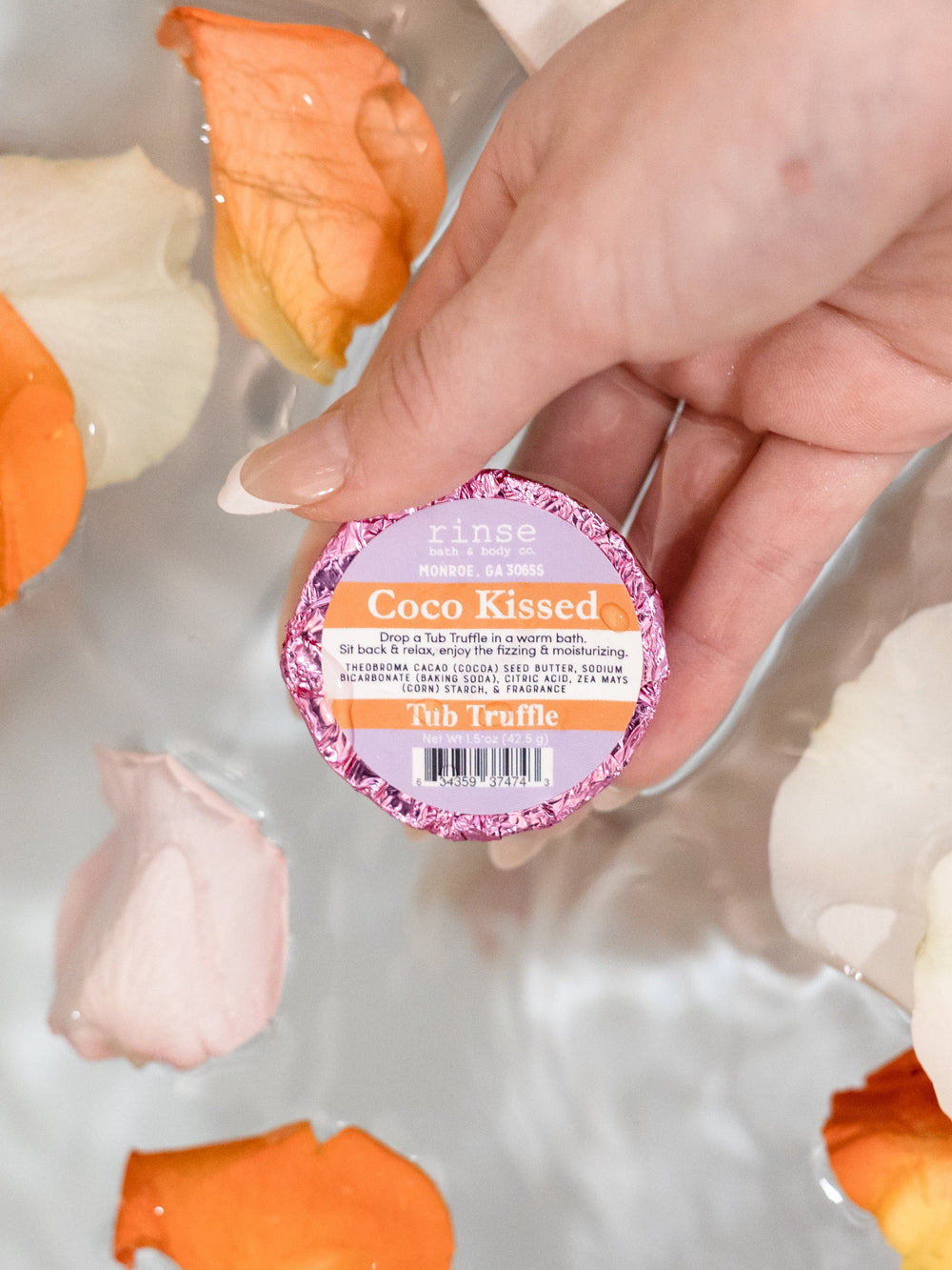 Hand holding pink-foil Coco Kissed Tub Truffle over bathwater — orange and white rose petals floating