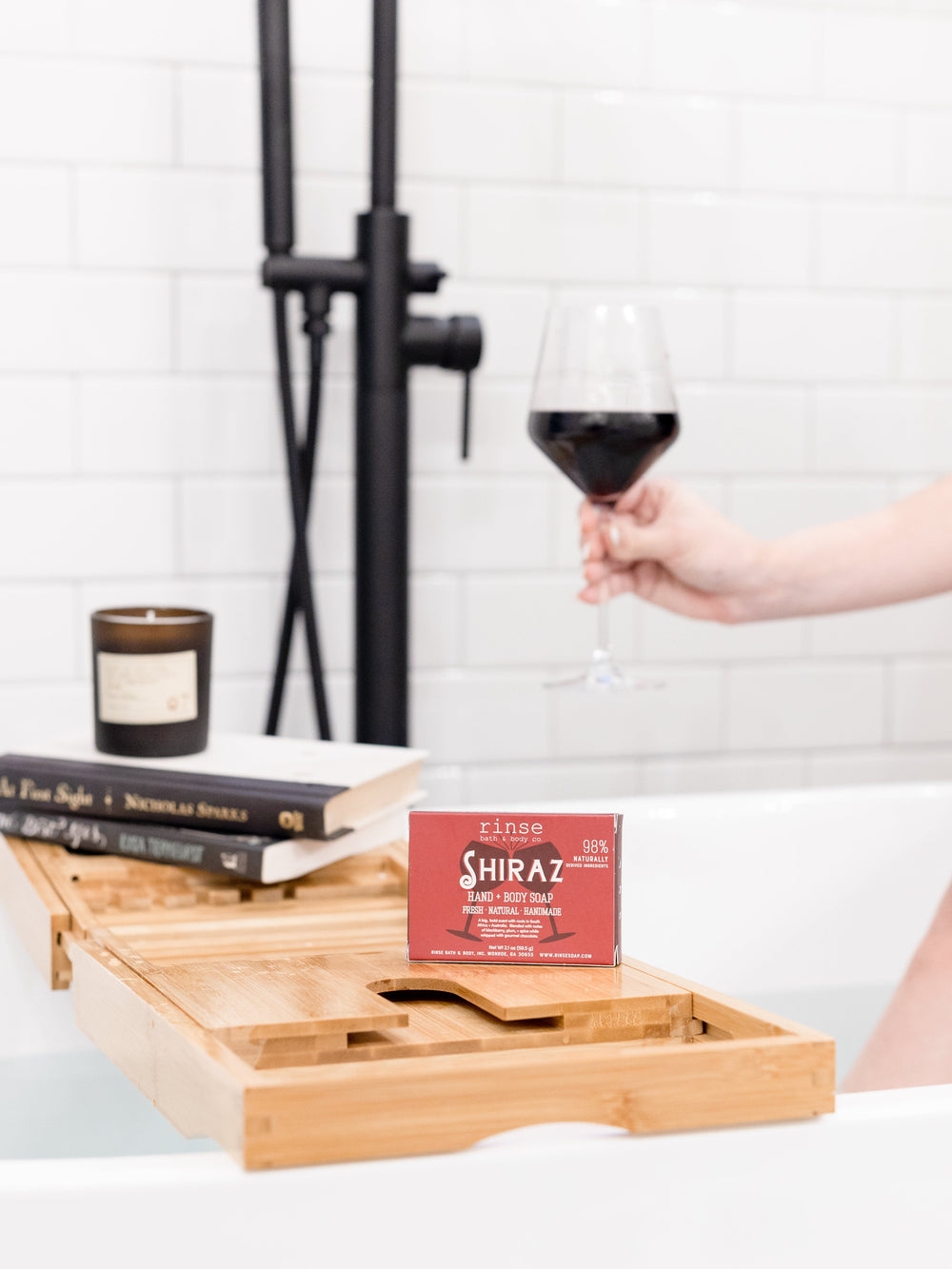 Shiraz soap on a bamboo bath tray with books and a candle — hand holding red wine over white tile tub