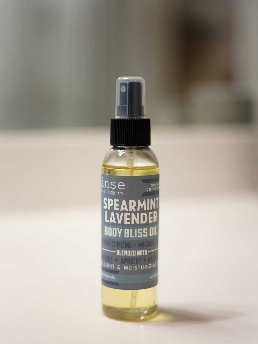 Spearmint Lavender Body Bliss Oil standing on reflective white — muted blue-gray label in soft spa bokeh