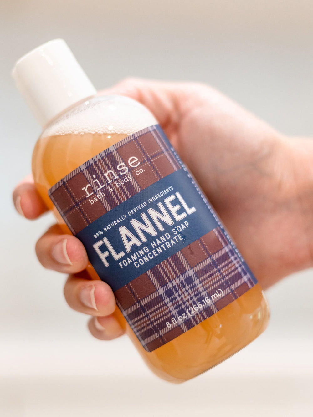 Hand tilting Flannel concentrate bottle toward camera — soft bubbles atop amber soap in bright indoor light