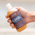 Hand tilting Flannel concentrate bottle toward camera — soft bubbles atop amber soap in bright indoor light