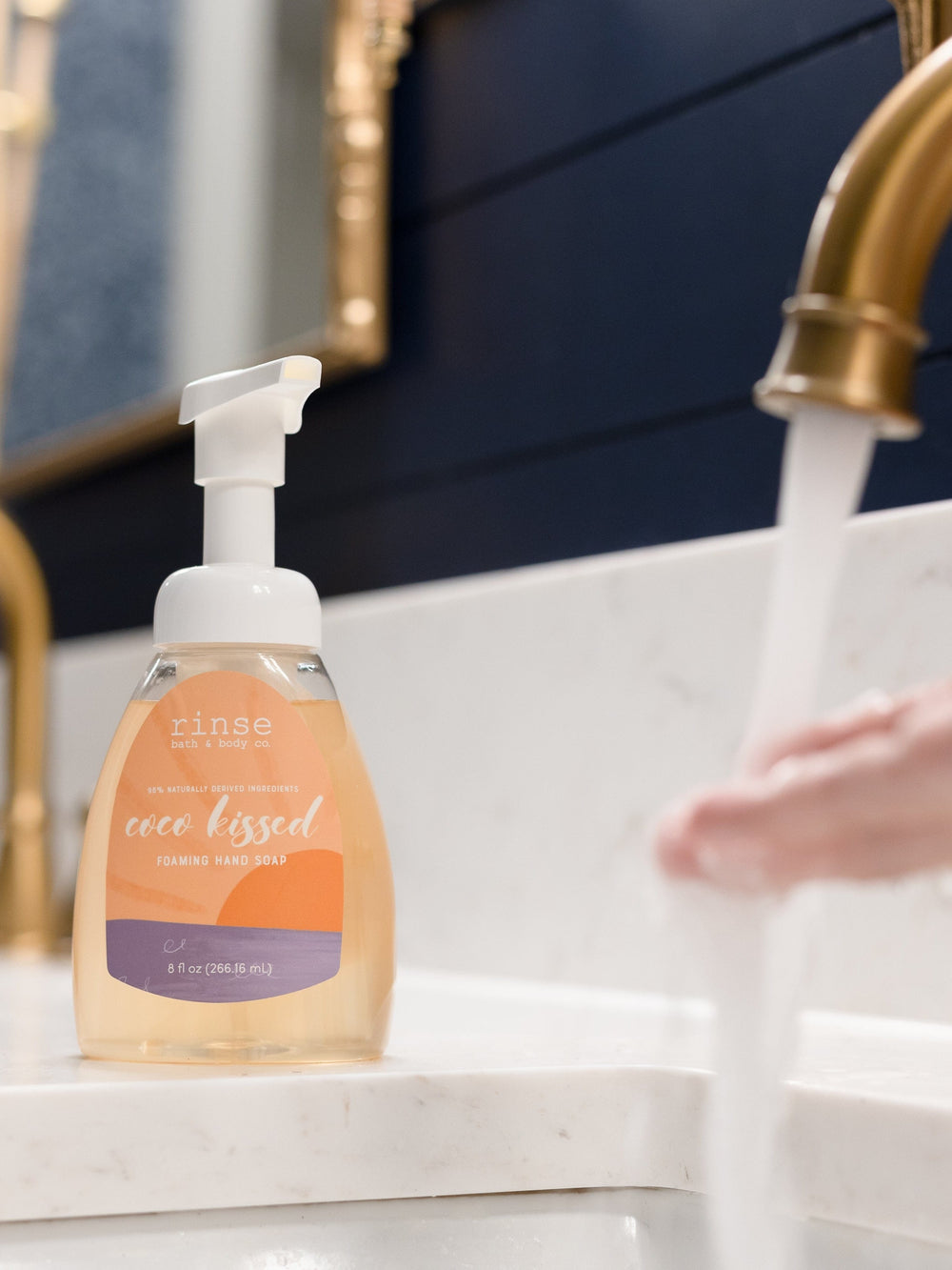 Coco Kissed foaming soap sits by a running gold faucet — navy paneling and marble vanity