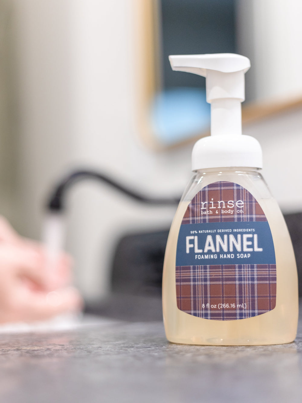 Flannel Foaming Hand Soap on a speckled gray counter while hands wash under a black faucet — soft bathroom blur