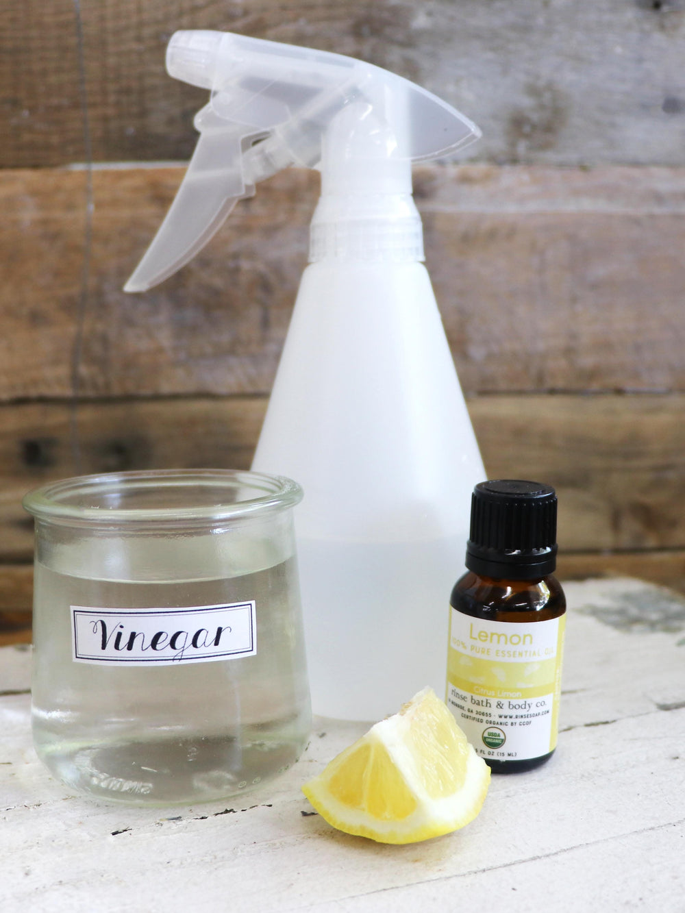 Lemon essential oil beside a juicy lemon wedge and vinegar jar — white wood table against dark rustic planks