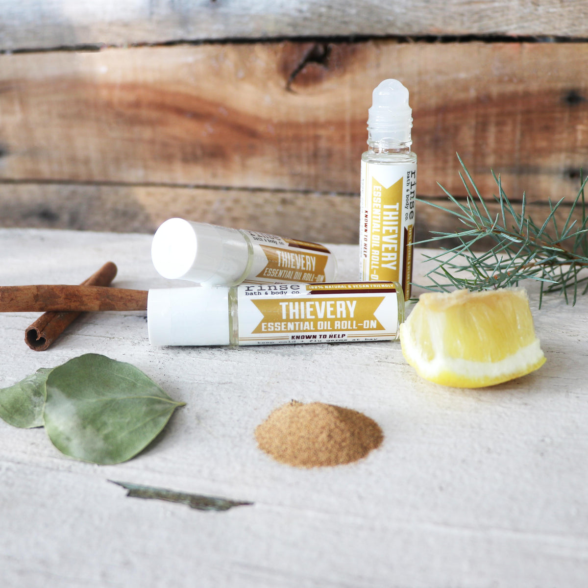 Three Thievery essential oil roll-on bottles arranged on a light fabric surface with cinnamon sticks, eucalyptus leaves, a lemon wedge, and a small pile of cloves, set against a rustic wooden plank background.