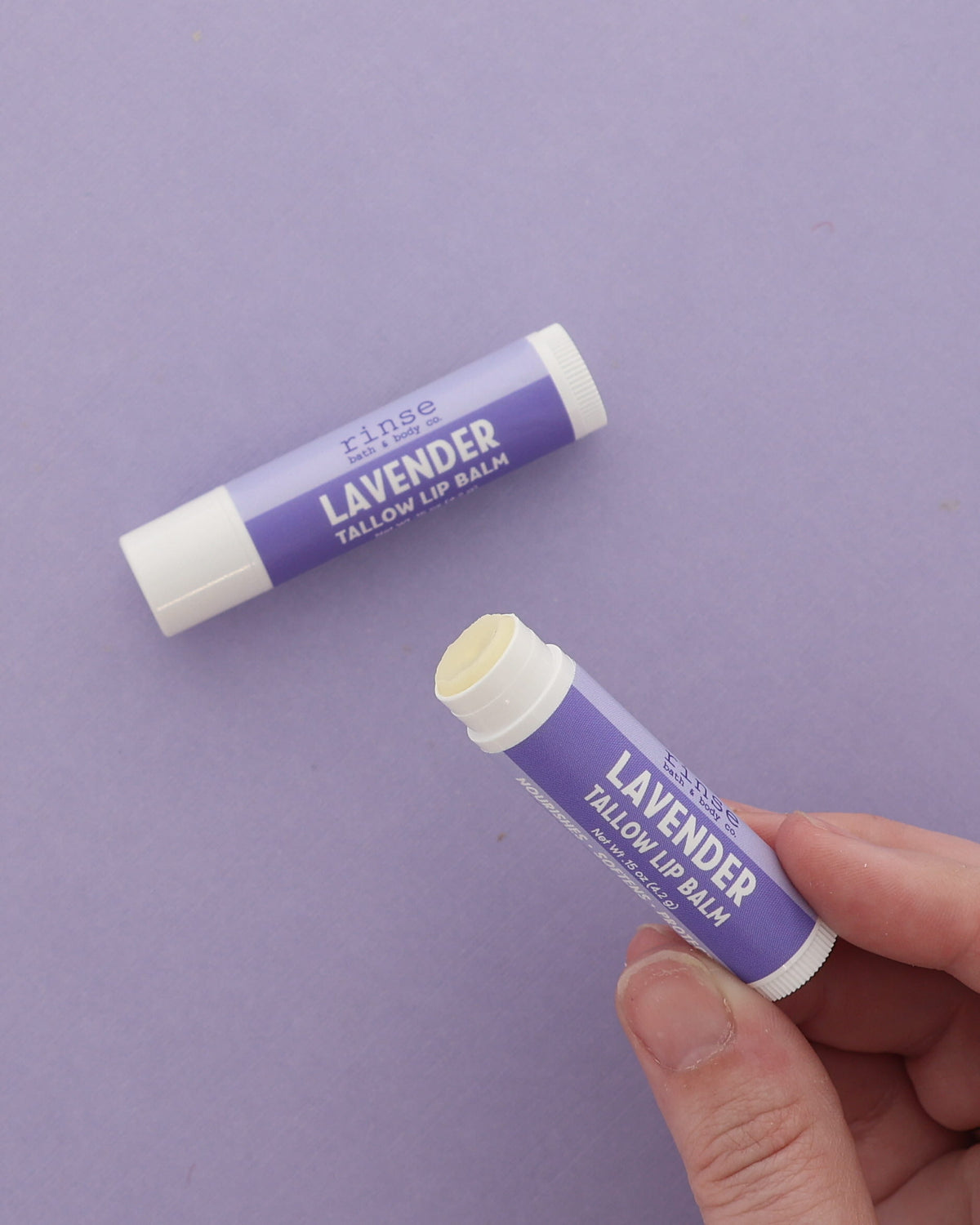 Hand holding an uncapped Lavender Tallow Lip Balm beside a capped tube on a plain lilac backdrop