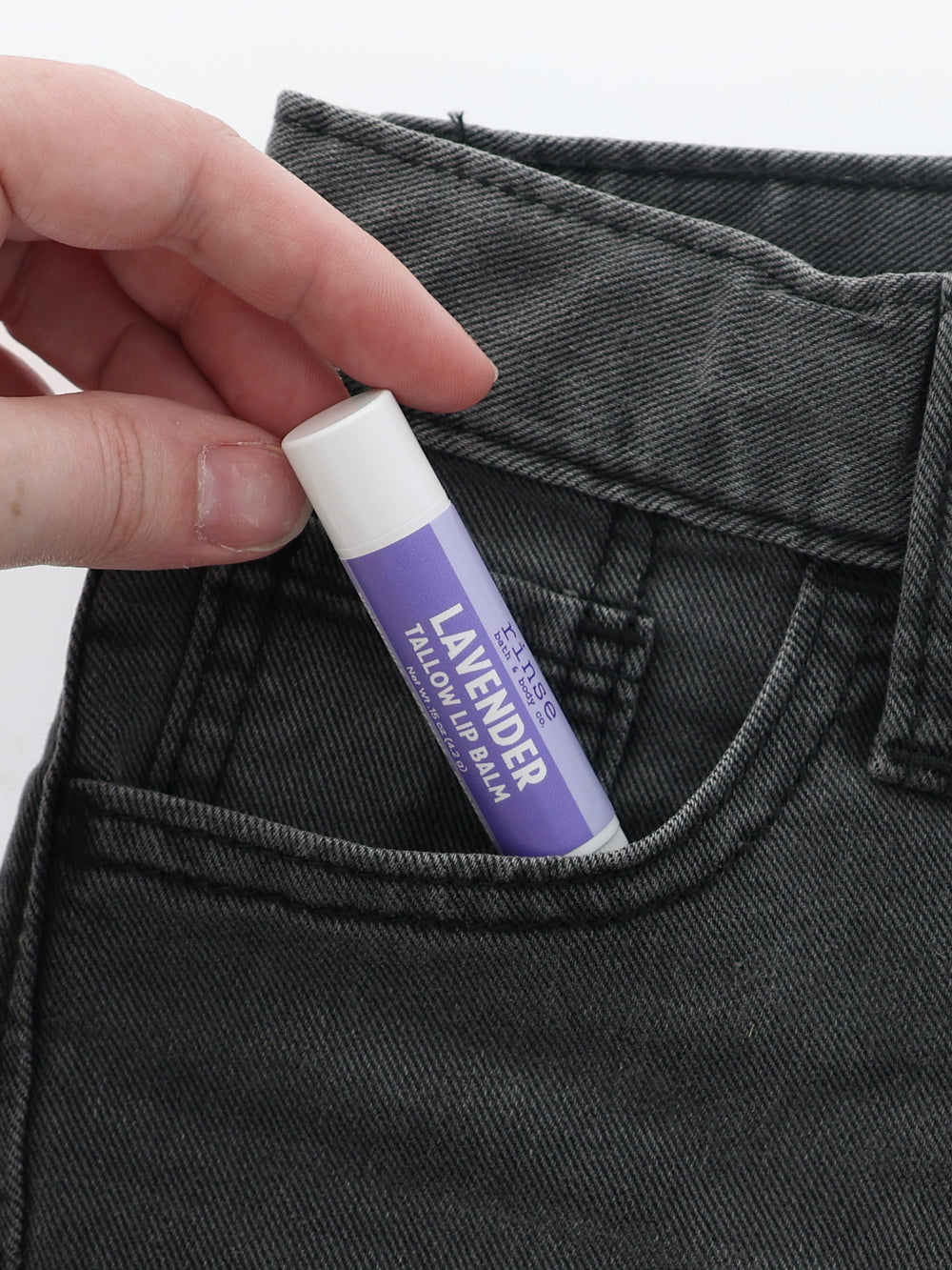 Lavender Tallow Lip Balm half-tucked into the front pocket of charcoal jeans, held between two fingers