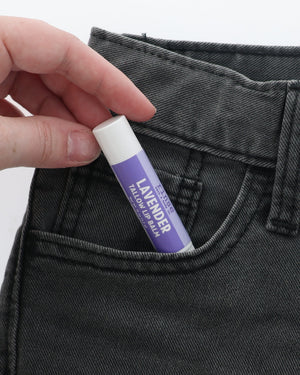 Lavender Tallow Lip Balm half-tucked into the front pocket of charcoal jeans, held between two fingers