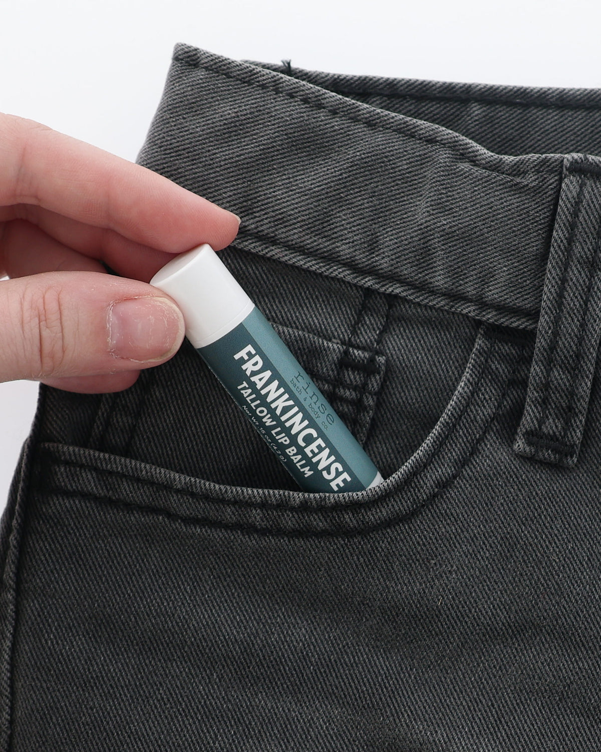 Frankincense Tallow Lip Balm half-tucked into the front pocket of charcoal jeans, held between two fingers