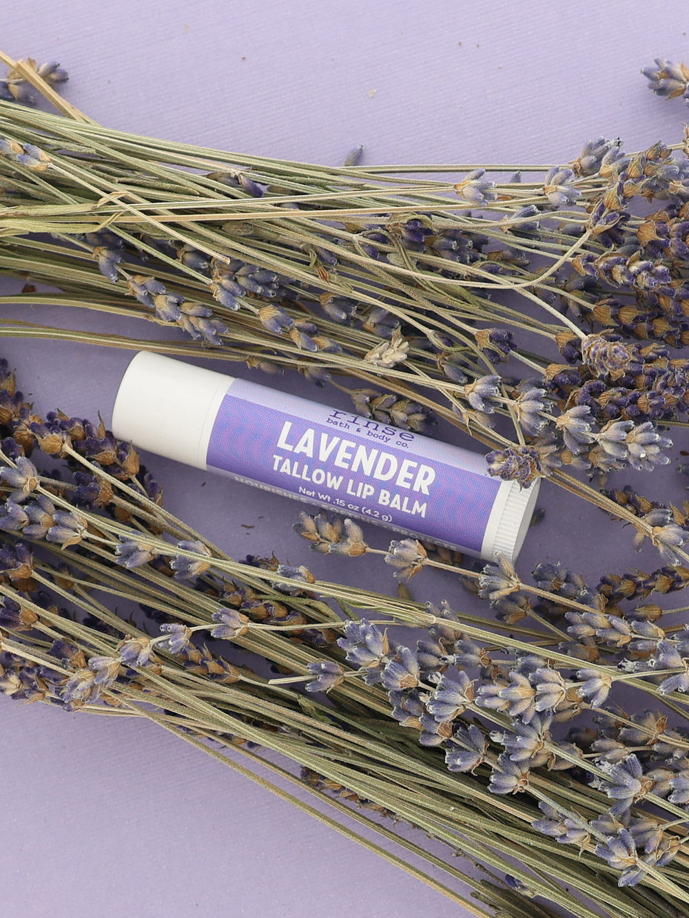 Lavender Tallow Lip Balm tube with purple label resting among dried lavender bundles on a lilac backdrop