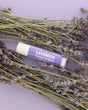 Lavender Tallow Lip Balm tube with purple label resting among dried lavender bundles on a lilac backdrop