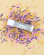 Vanilla Tallow Lip Balm tube with grey label lying on pink, purple and gold sprinkles on a butter-yellow backdrop