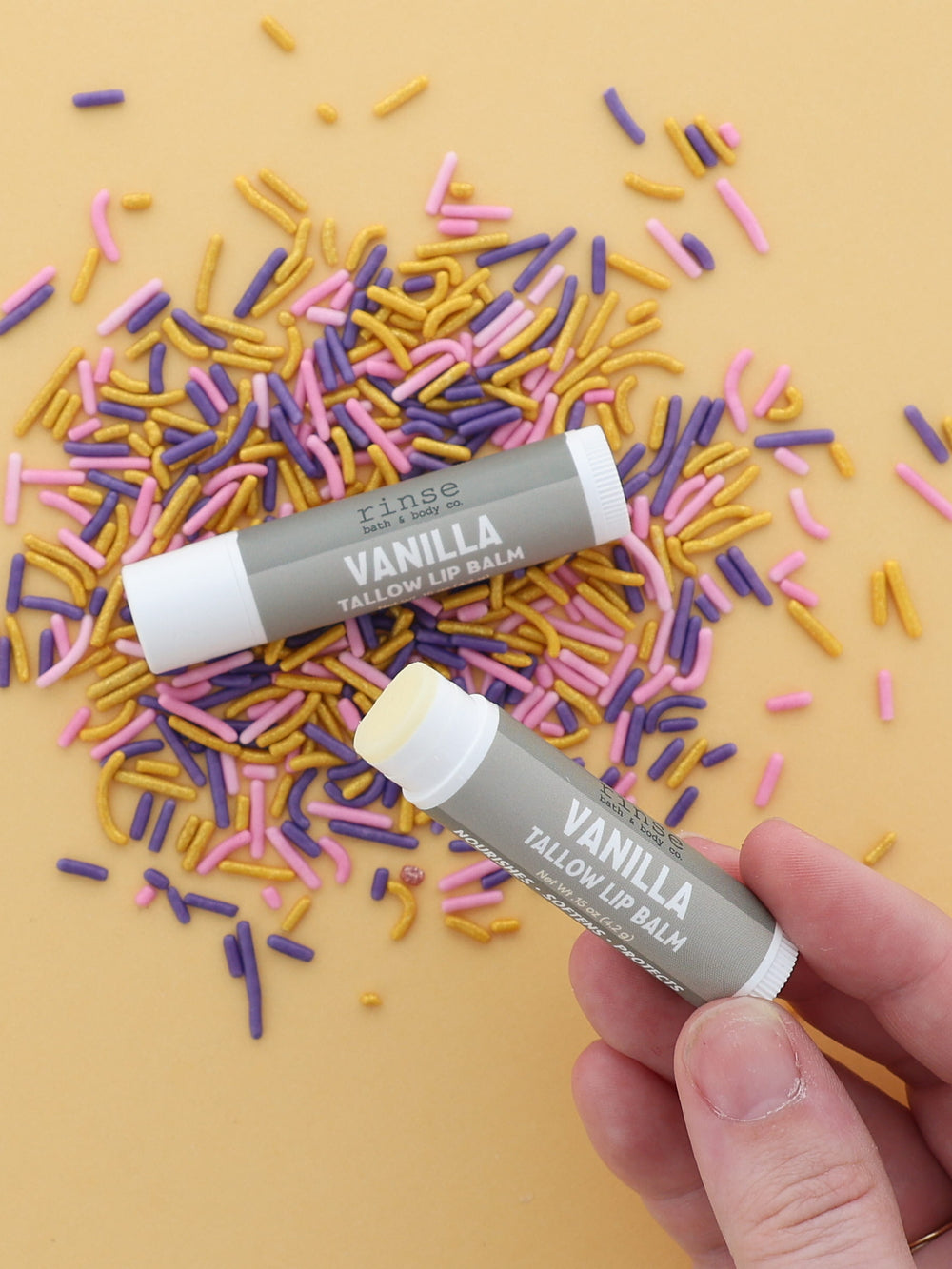 Hand holding an uncapped Vanilla Tallow Lip Balm beside a capped tube on pink, purple and gold sprinkles