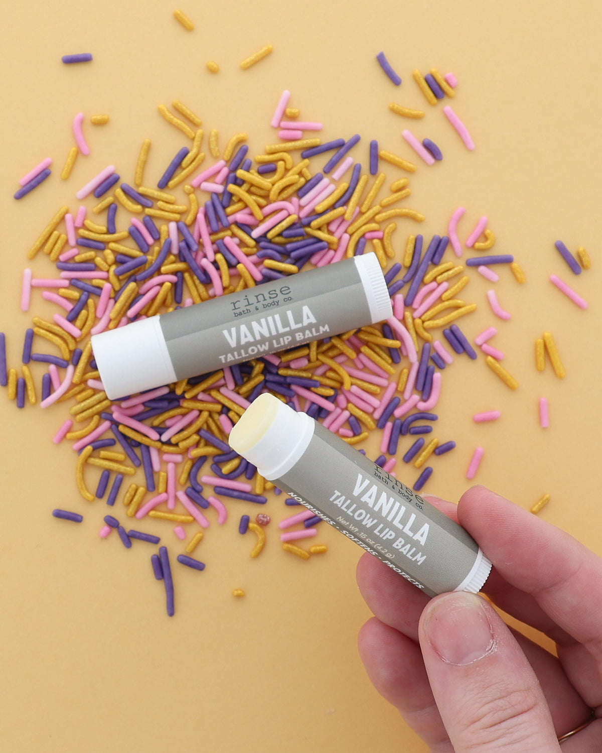 Hand holding an uncapped Vanilla Tallow Lip Balm beside a capped tube on pink, purple and gold sprinkles
