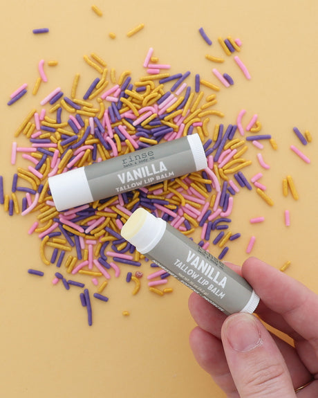 Hand holding an uncapped Vanilla Tallow Lip Balm beside a capped tube on pink, purple and gold sprinkles