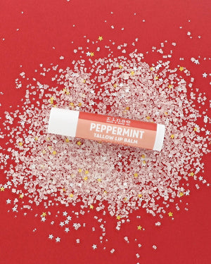 Peppermint Tallow Lip Balm tube with coral-red label lying on pink-white crystals and tiny gold and silver stars on red