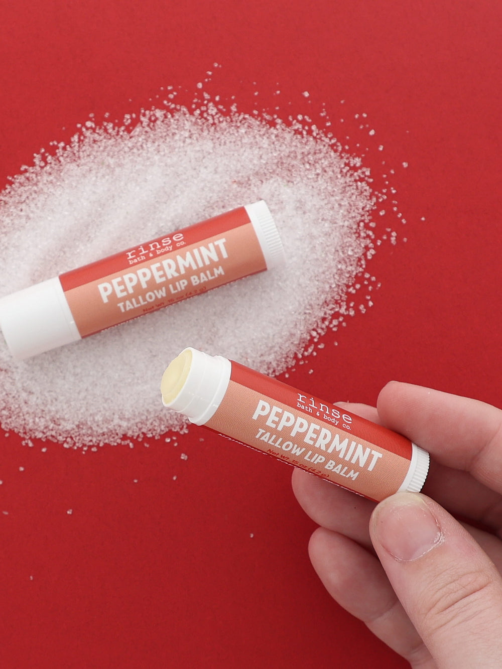 Hand holding an uncapped Peppermint Tallow Lip Balm beside a capped tube on a pile of white crystals on red