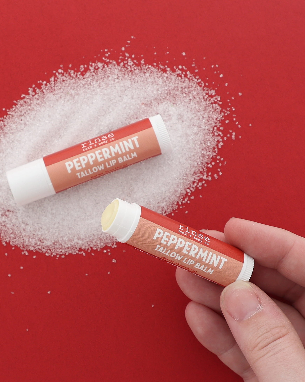 Hand holding an uncapped Peppermint Tallow Lip Balm beside a capped tube on a pile of white crystals on red