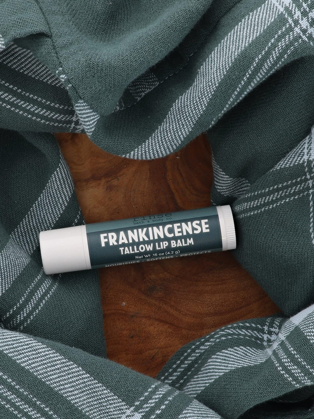 Frankincense Tallow Lip Balm tube with deep teal label lying on a wooden board framed by green plaid flannel