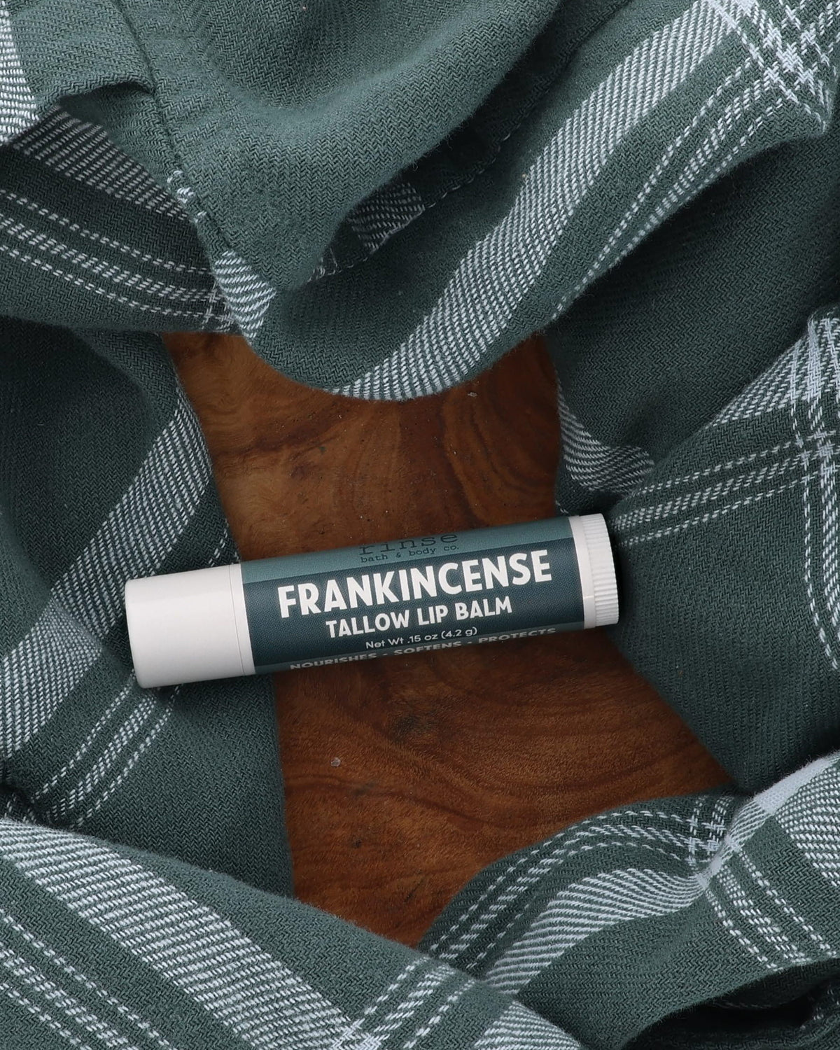 Frankincense Tallow Lip Balm tube with deep teal label lying on a wooden board framed by green plaid flannel