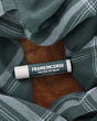 Frankincense Tallow Lip Balm tube with deep teal label lying on a wooden board framed by green plaid flannel