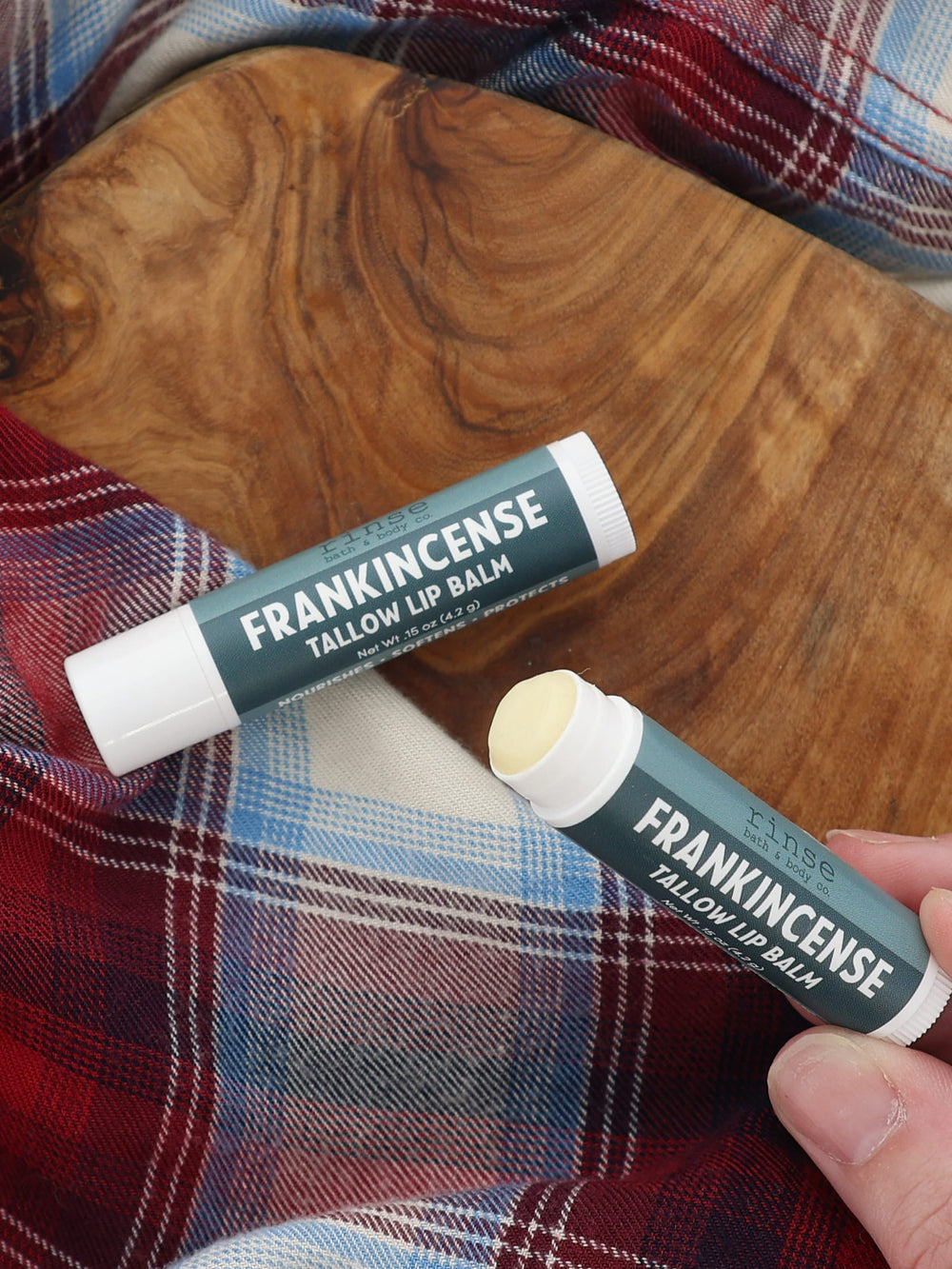 Hand holding an uncapped Frankincense Tallow Lip Balm beside a capped tube on a wooden board and red-and-blue plaid