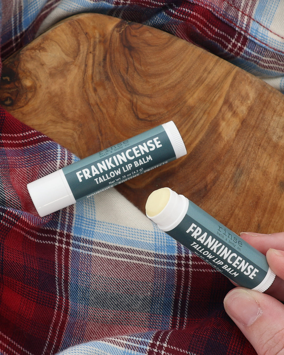 Hand holding an uncapped Frankincense Tallow Lip Balm beside a capped tube on a wooden board and red-and-blue plaid