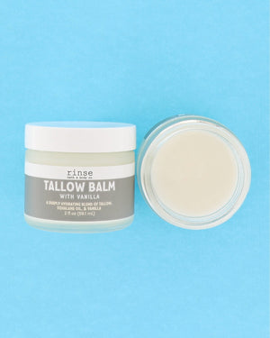 A jar of Rinse Tallow Balm with vanilla next to its open lid, showing the creamy off-white balm inside, set against a solid light blue background.
