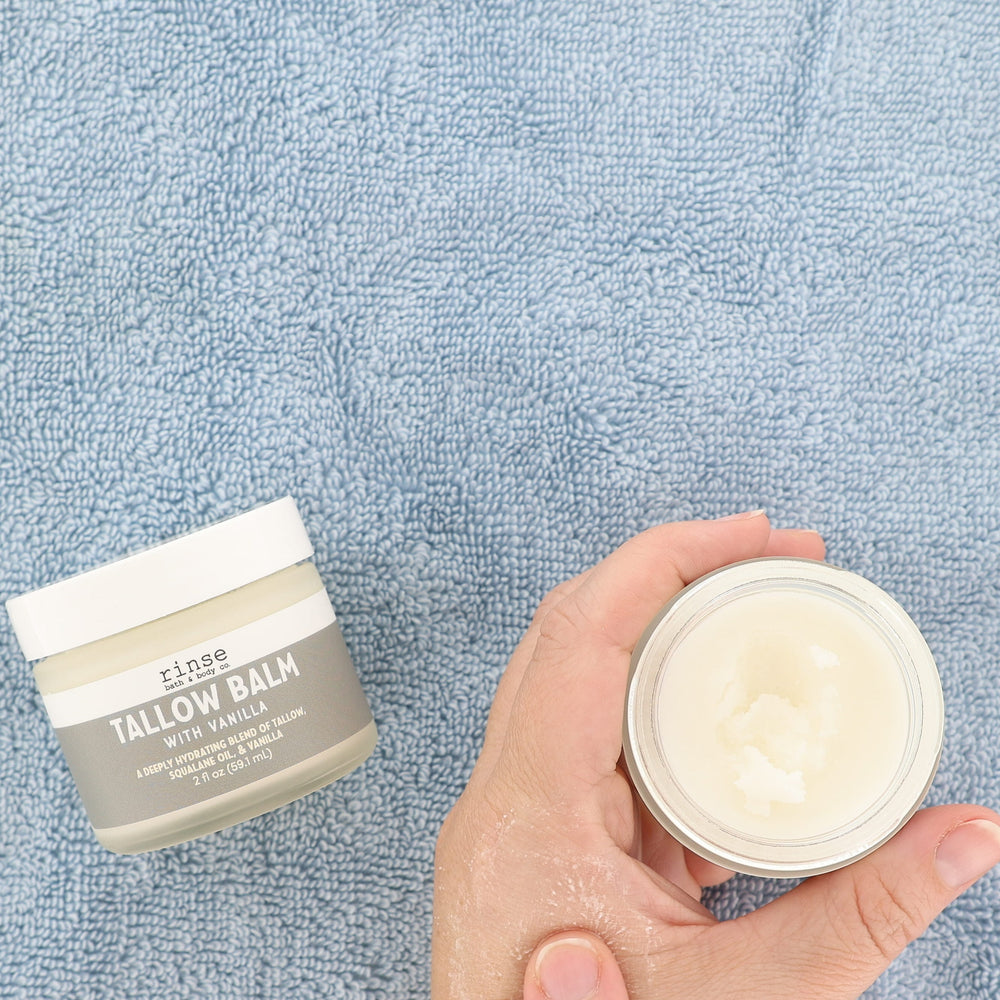 A hand holds an open jar of white balm next to a closed jar labeled 'close TALLOW BALM WITH VANILLA' on a textured light blue towel background.