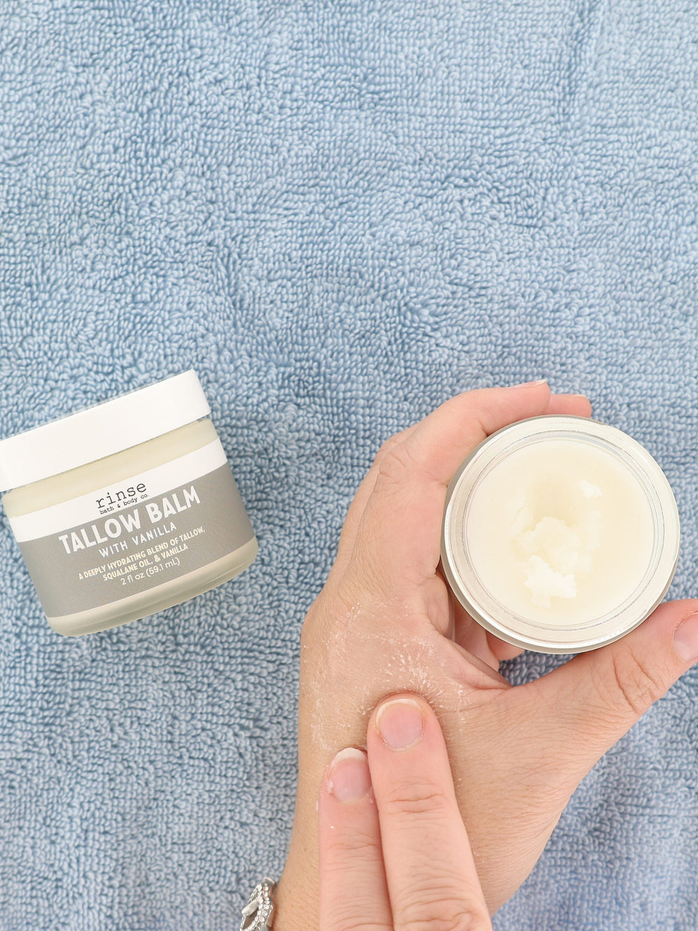 Hands rubbing creamy Tallow Balm with Vanilla onto the back of a hand — open jar on a soft blue towel
