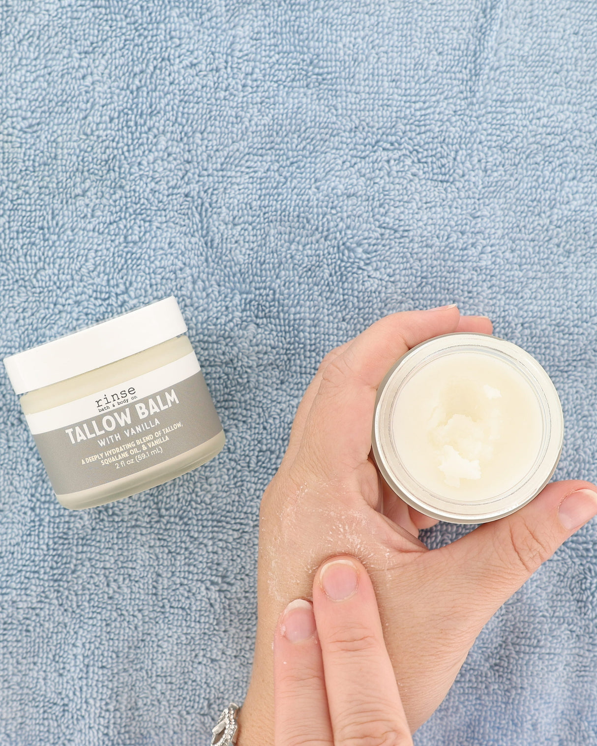 A hand holds an open jar of white balm next to a closed jar labeled 'close TALLOW BALM WITH VANILLA' on a textured light blue towel background.