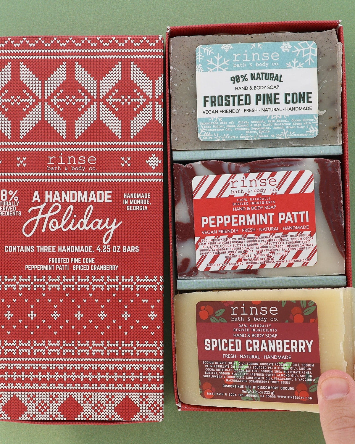 Open Handmade Holiday box on green holding Frosted Pine Cone, Peppermint Patti, and Spiced Cranberry — Fair Isle lid beside three festive bars