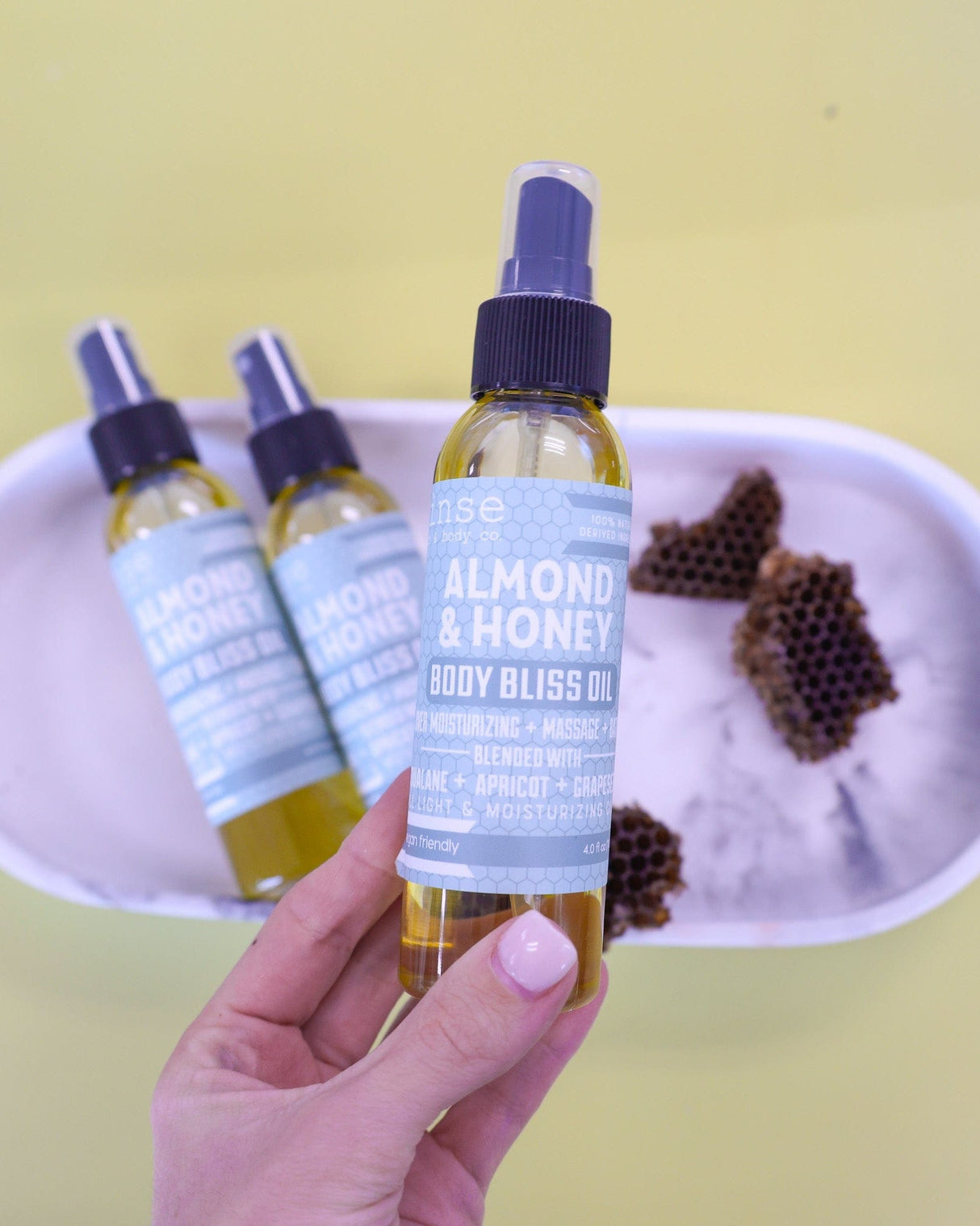 A hand lifting Almond & Honey Body Bliss oil above a lavender tray set with raw honeycomb — warm almond and honey in a silky oil that sinks straight in. Rinse Bath & Body Co.