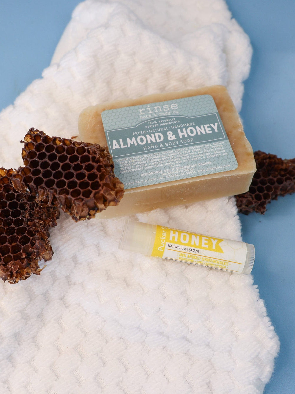 Almond & Honey bar and Honey Pucker Stick resting on a waffle towel with honeycomb — spa-calm blue surface in soft light