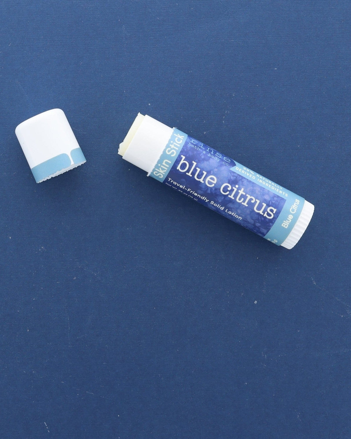 A Blue Citrus Skin Stick lying on a deep blue surface with its cap off, the twist-up solid lotion showing at the top — travel-friendly solid lotion in a pocket-sized tube. Rinse Bath & Body Co.