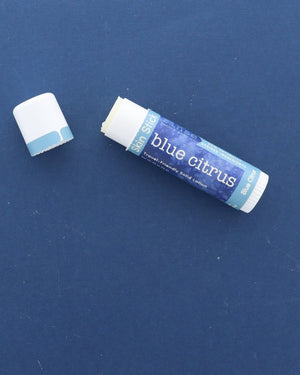 A Blue Citrus Skin Stick lying on a deep blue surface with its cap off, the twist-up solid lotion showing at the top — travel-friendly solid lotion in a pocket-sized tube. Rinse Bath & Body Co.