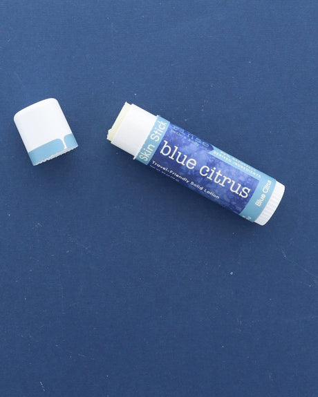 A Blue Citrus Skin Stick lying on a deep blue surface with its cap off, the twist-up solid lotion showing at the top — travel-friendly solid lotion in a pocket-sized tube. Rinse Bath & Body Co.