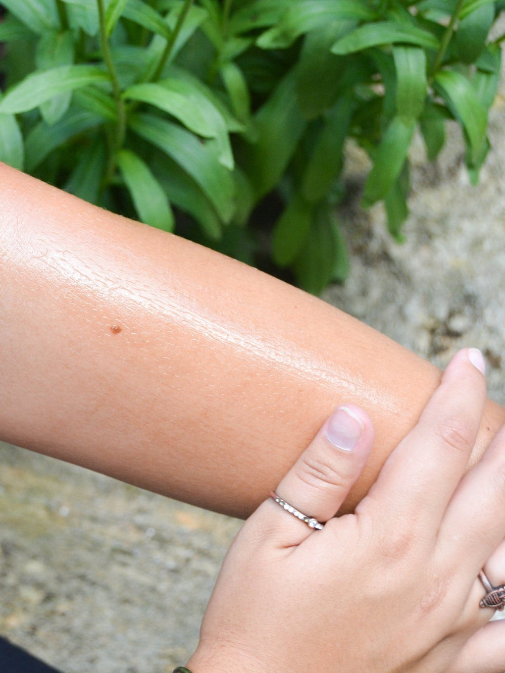 Hand smoothing glossy Cabernet oil along a forearm outdoors — dewy skin against leafy garden light
