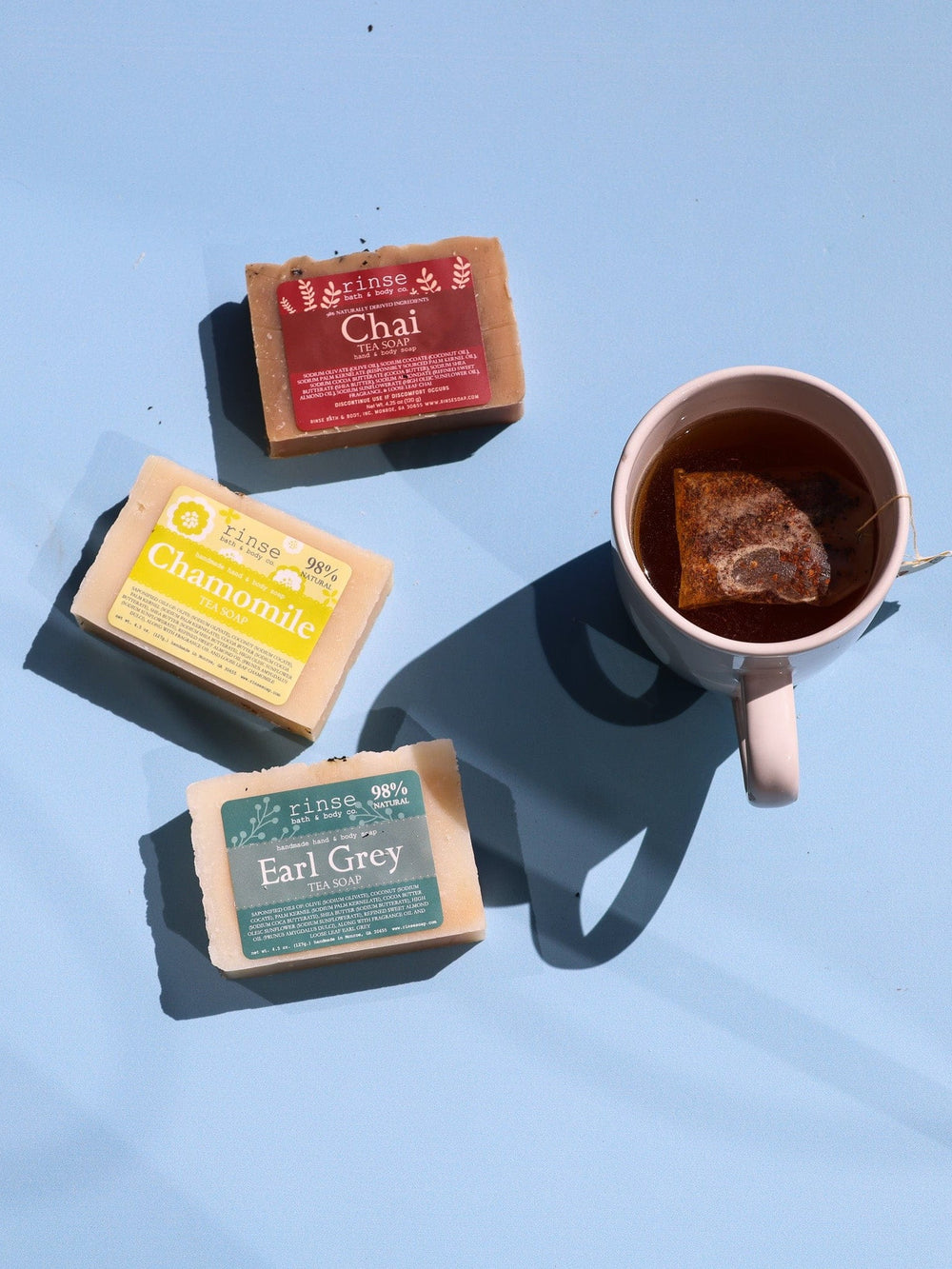 Chai Chamomile and Earl Grey tea soaps circle a rose mug — sharp sun shadows on pale blue