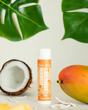 Coconut Mango Skin Stick standing between halved coconut and whole mango amid green leaves — tropical solid lotion