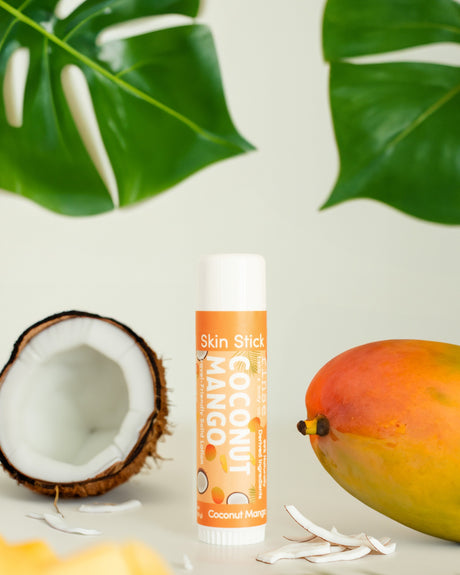 Coconut Mango Skin Stick standing between halved coconut and whole mango amid green leaves — tropical solid lotion