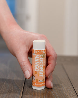 Hand holding open Coconut Mango Skin Stick solid lotion on dark wood — travel lotion ready for dry spots
