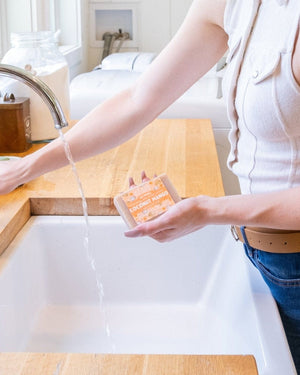 Hands holding Coconut Mango bar soap under a running chrome faucet — white farmhouse sink, everyday shower cleanse ritual