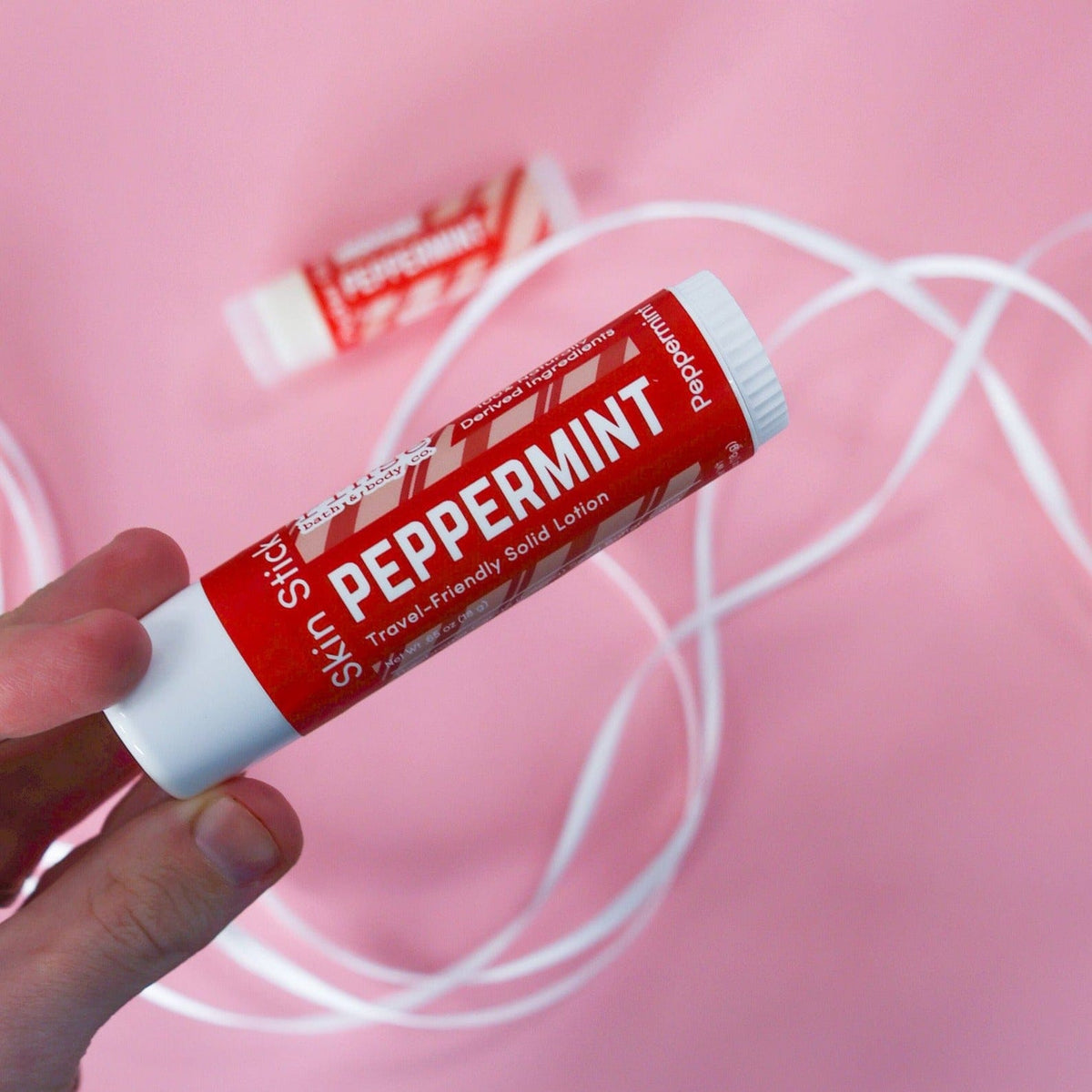 Hand holding Peppermint Skin Stick against bubblegum pink — solid lotion tube in lifestyle light