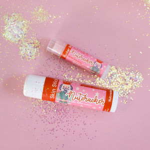 Nutcracker Skin Stick held in soft studio light — pastel pink ground with glitter flecks, solid lotion twist-up in use