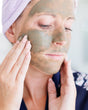 A woman smoothing green Dead Sea mud mask across her cheeks with both hands, a lavender headband holding her hair back — a detoxifying clay treatment for the week your skin has had. Rinse Bath & Body Co.