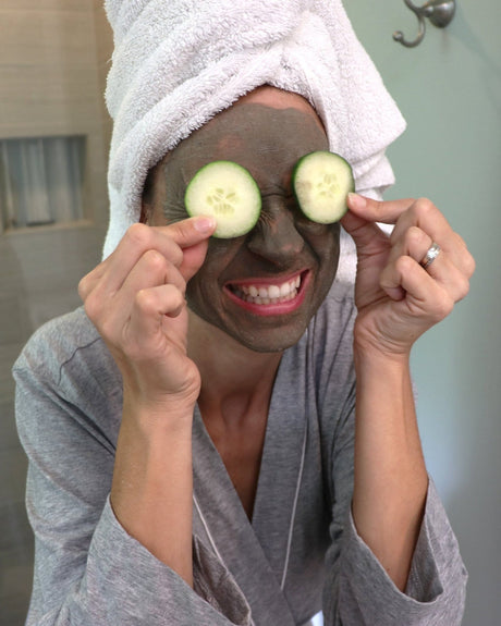 A woman in a robe and towel turban holding cucumber slices over her eyes, Dead Sea mud mask dried to a matte finish across her face — an unhurried at-home spa afternoon. Rinse Bath & Body Co.