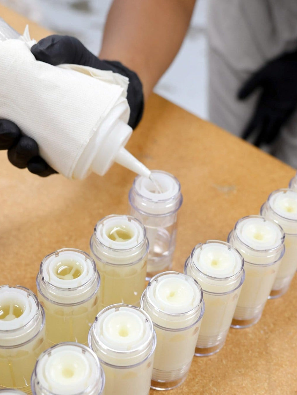 Gloved hands pouring warm DeOdor into rows of empty tubes — wooden workbench lined with clear sticks