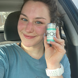 Smiling woman holds clear Original deOdor stick beside her face in the car — teal sweatshirt and soft daylight